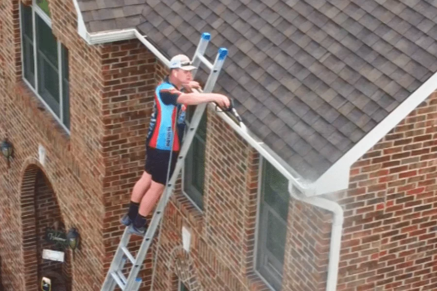 A man precariously stands on a ladder, using a soft wash technique to clean the outside of gutters on a brick house. He holds a pressure washer wand while leaning against the roof.