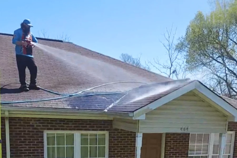 Man soft washing a shingle roof with a pressure washer. He's wearing a blue hat and safety vest, standing on the roof of a brick house, spraying away dirt and algae.