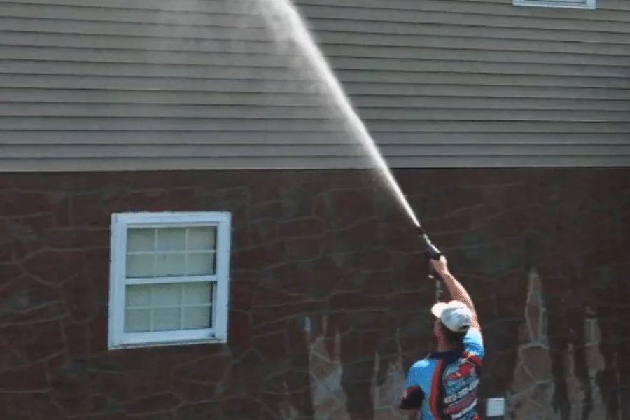 A worker uses a power washer to soft wash siding on a two-story house with stone and vinyl exterior. The stream of water reaches the upper level.