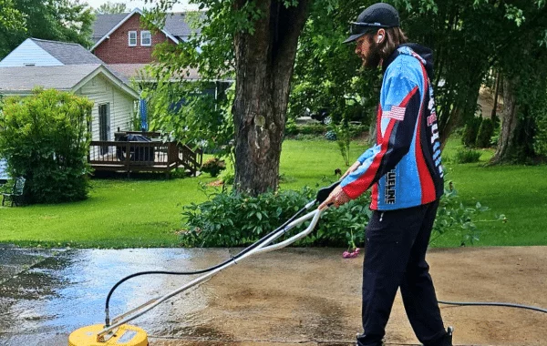 Man pressure washing a concrete drive-thru area with a surface cleaner attachment. He wears a blue and red jacket and a baseball cap. Green lawn, trees, and houses are in the background.