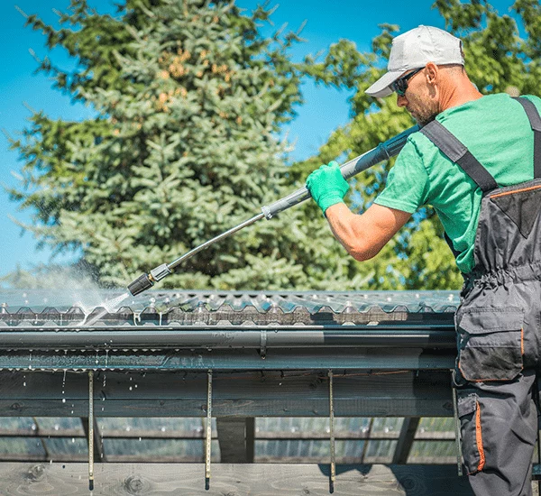 Man soft washing painted gutters with a pressure washer. He wears safety glasses, gloves, and overalls, with a green tree in the background.