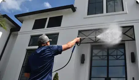 Man power washing the exterior of a modern white house. The pressure washing spray is visible against the clean facade, highlighting a home maintenance service.
