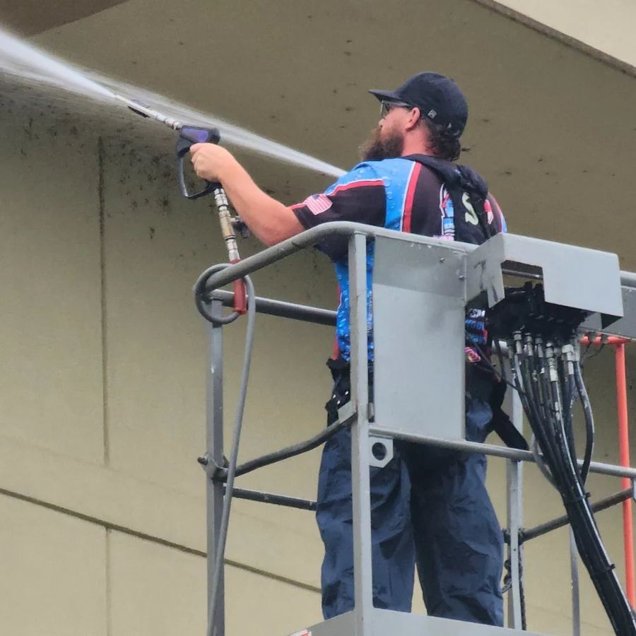 A worker on a lift uses a pressure washer to clean a painted wall. This soft washing technique can safely clean surfaces without damage.