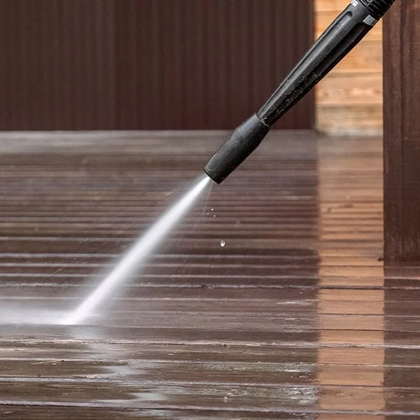 Power washing a wooden deck. A stream of water blasts away dirt and grime, revealing the clean wood underneath. The dark wood siding is visible in the background.
