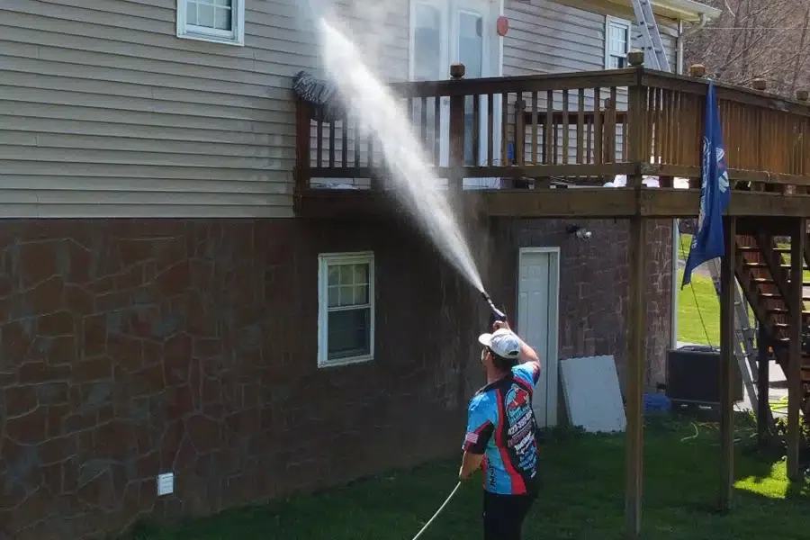 A man soft washing vinyl siding on a two-story house. He uses a pressure washer to gently clean the siding, aiming the spray upwards. The house has a stone foundation and a wooden deck.