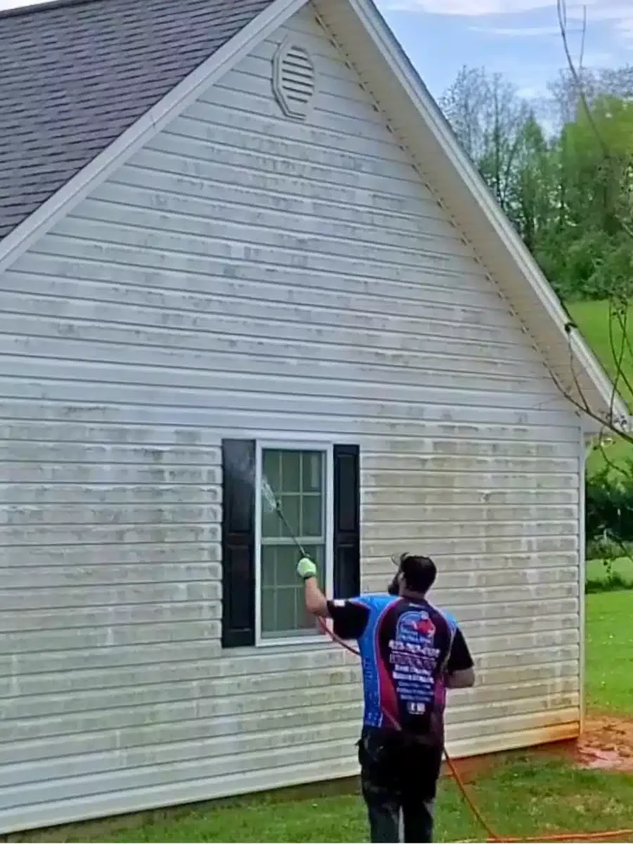 A man soft washing vinyl siding, using a long wand to spray the exterior of a house covered in green algae and dirt. The house has black shutters and is surrounded by a green lawn.