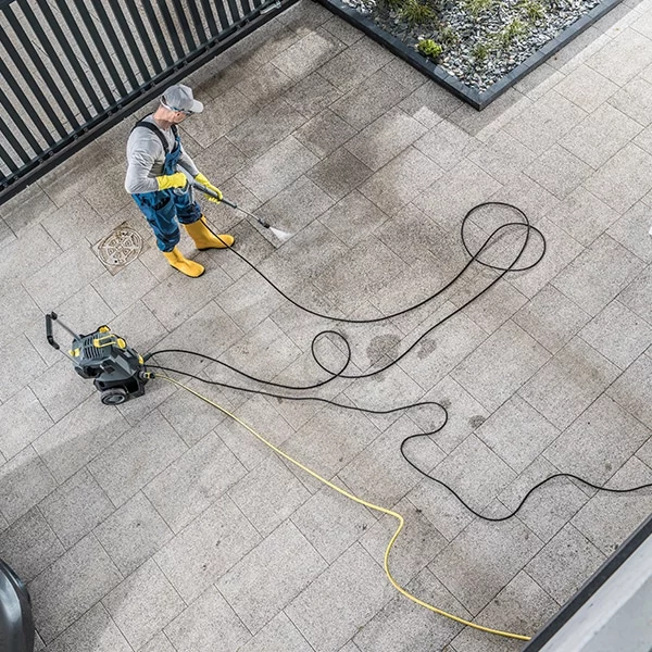 A man power washing a stone patio with a Kärcher pressure washer. He wears yellow boots and gloves, using a wand attachment to clean the stone surface. Black and yellow hoses snake across the patio.